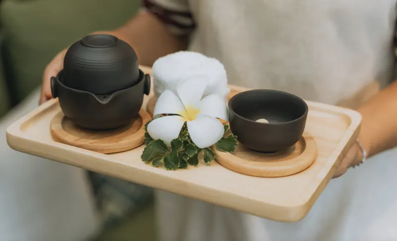 Server holding wooden tray with black teapot, cups, white frangipani flowers, and greens