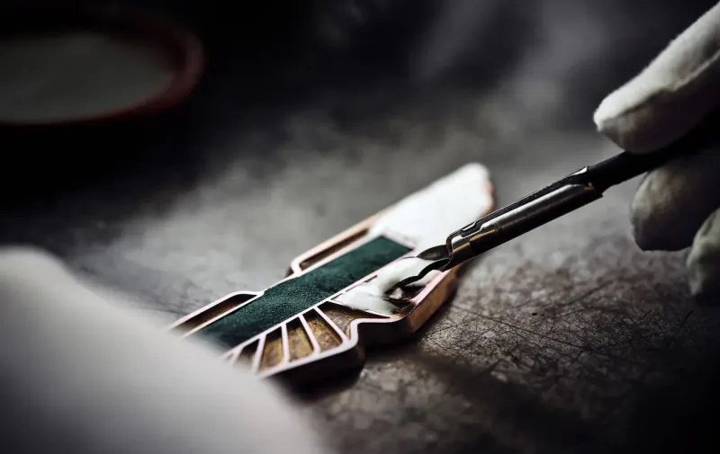 Gloved hand using tweezers to apply green enamel to Aston Martin wings logo on workbench