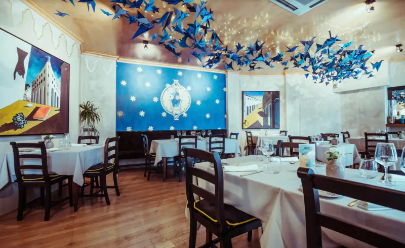Elegant Italian restaurant interior with blue paper birds hanging from ceiling, white tables set with glassware, and abstract wall art.