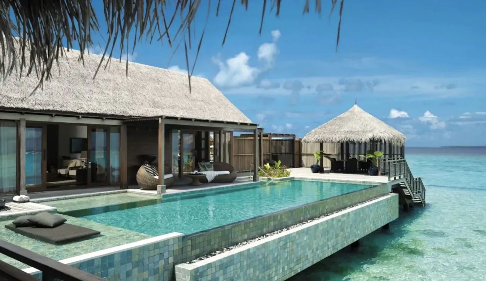 Luxury overwater villa with infinity pool extending over turquoise lagoon, palm-fringed thatched roofs.