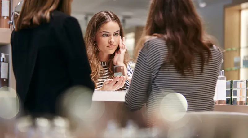 Two women at cosmetics counter: one facing mirror, touching cheek thoughtfully; beauty products on shelves.