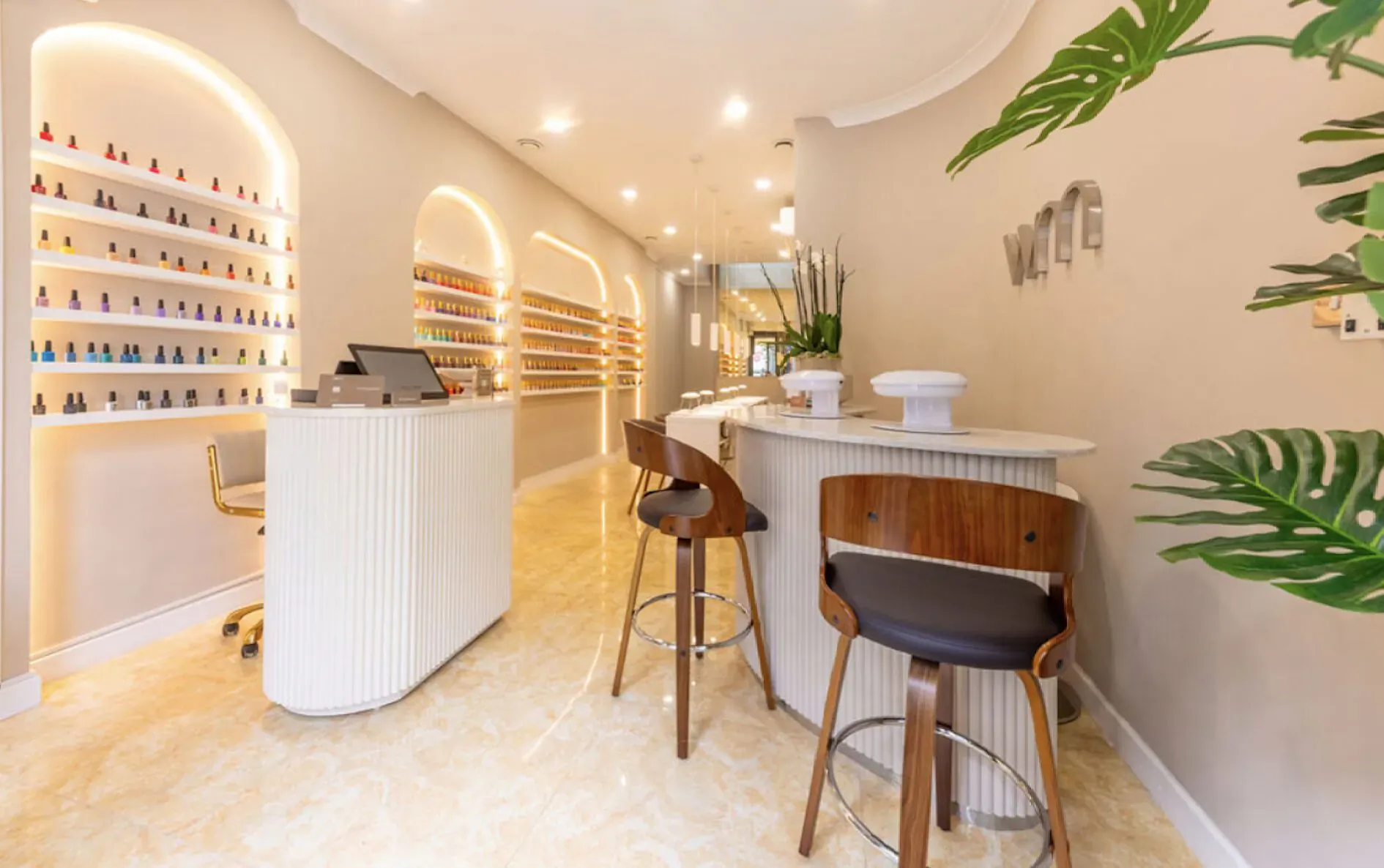 Modern nail salon interior with curved white reception, wooden stools, nail polish shelves, and plants in Fitzrovia spa.