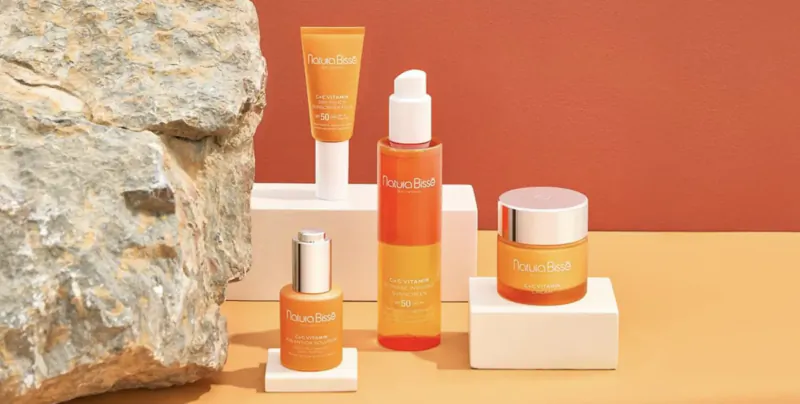 Natura Bissé skincare products on pedestals beside a large rock: orange tube, serum, oil, and cream jar.