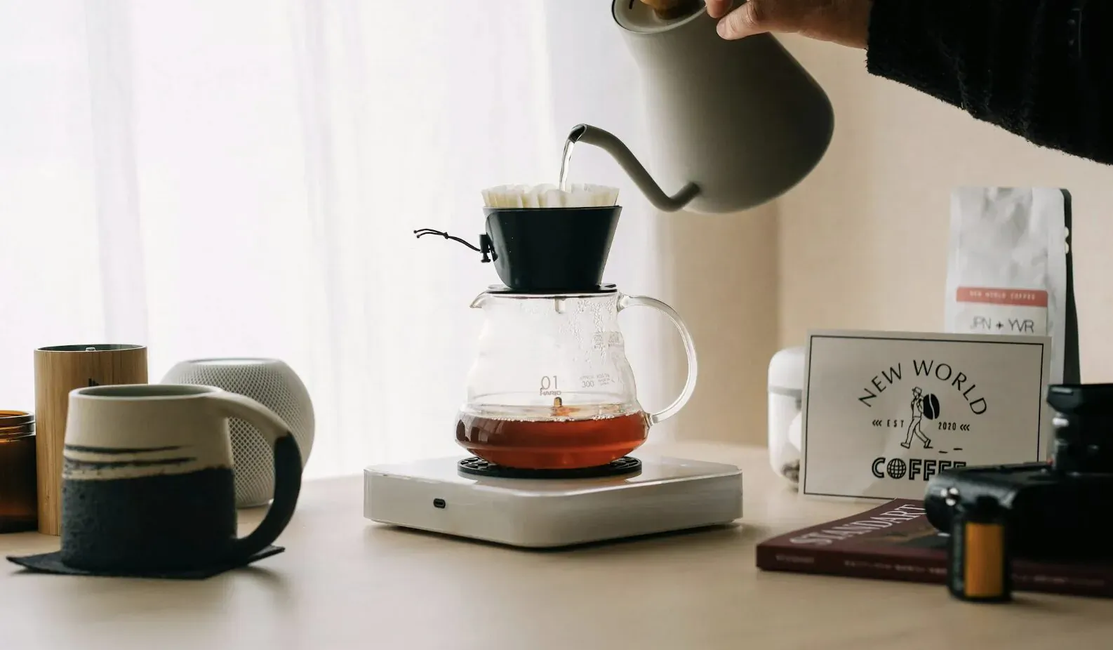 Hand pouring water from gooseneck kettle over coffee grounds in glass pour-over dripper on white base, 'New World' sign, mugs, and coffee bag nearby.