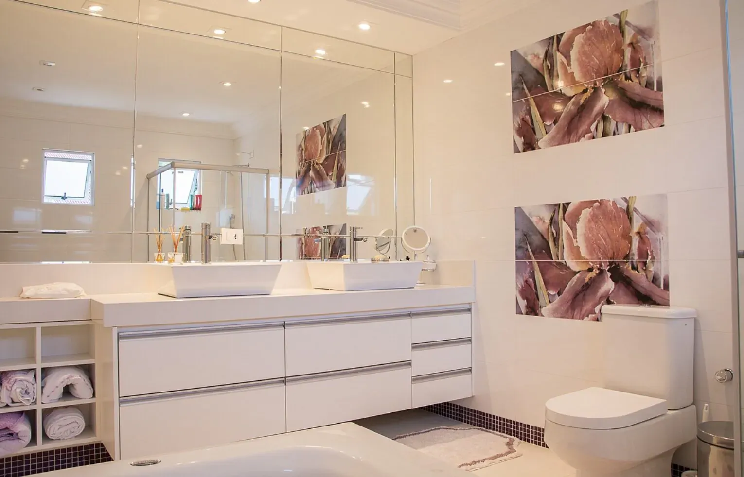 Modern white bathroom with double sinks, cabinets, toilet, tub, and floral wall art.