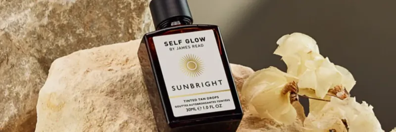 Self Glow Sunbright serum bottle on beige rocks with yellow orchids, labeled 'Self Glow Sunbright 30ml'.