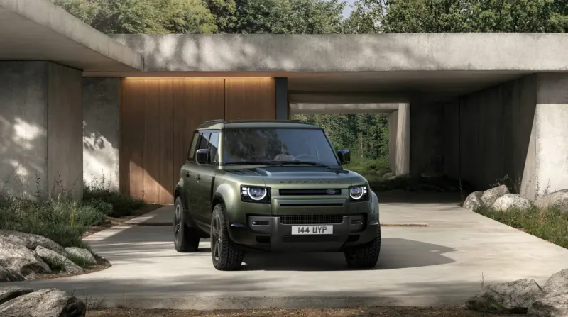 Green Land Rover Defender parked in modern concrete garage with greenery, license plate 'DEFENDER'.