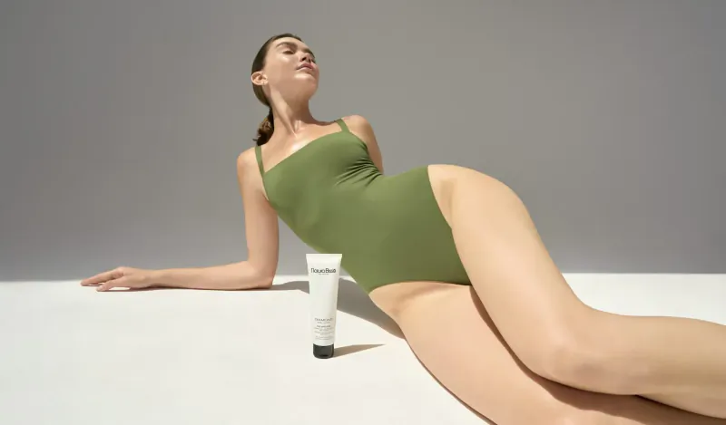 Model in green one-piece swimsuit reclines on white surface next to Natura Bissé Lipo-Gel Sculpting tube.