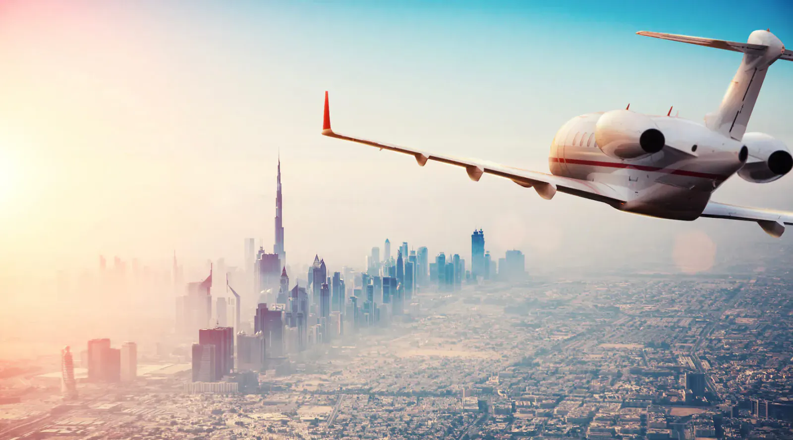 White private jet flying over hazy Dubai skyline with Burj Khalifa at golden sunset.