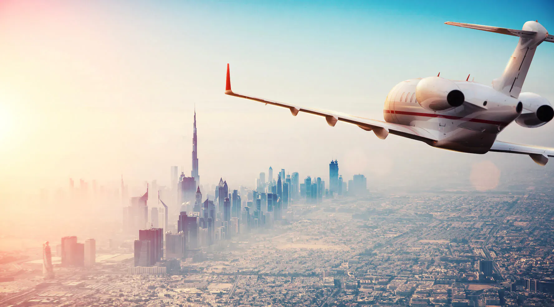 White private jet flying over hazy Dubai skyline with Burj Khalifa at golden sunset.