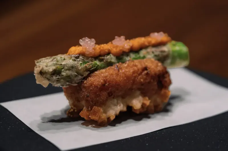 Tempura asparagus croquette sushi topped with orange paste and pink seaweed flakes on white paper