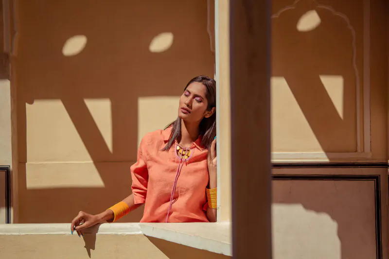 Indian woman in orange blouse and necklace leans pensively on windowsill in sunlit Rajasthani architecture