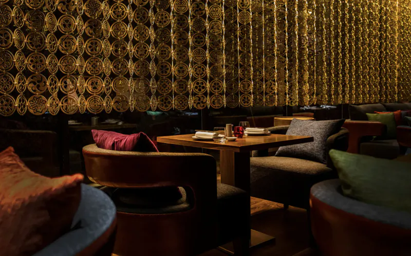 Myazú Restaurant’s vibrant interior with ornate gold curtains, plush colorful cushions on wooden chairs, and candlelit table.