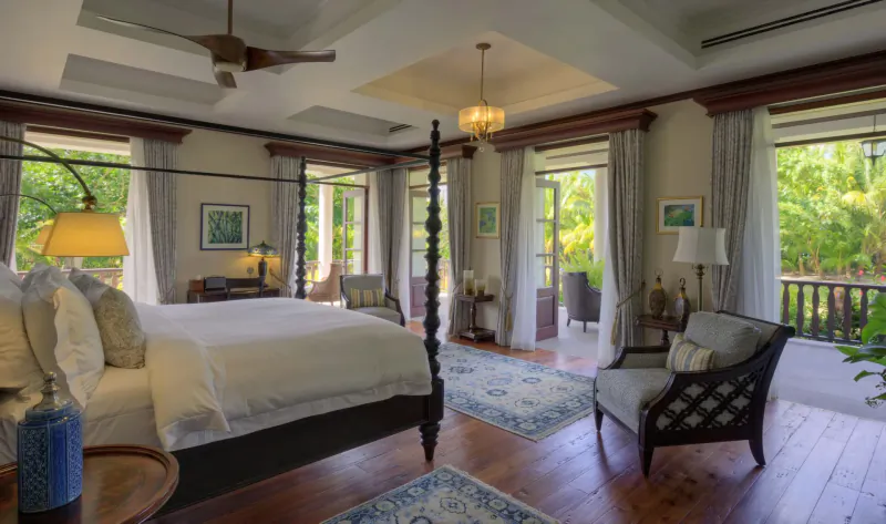 Elegant four-poster bedroom in La Cigale Estate, Seychelles, with balcony views of tropical greenery, ceiling fan, and lamps.