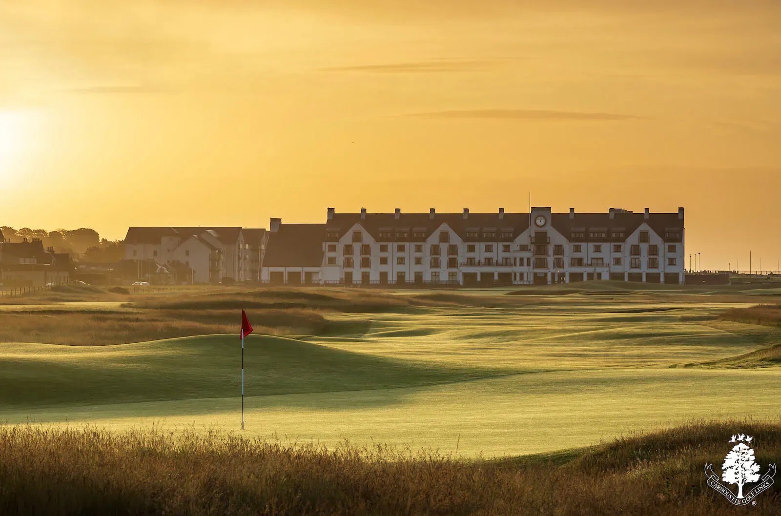 Carnoustie Golf Links: white clubhouse and green fairway with red flag at sunset in Scotland