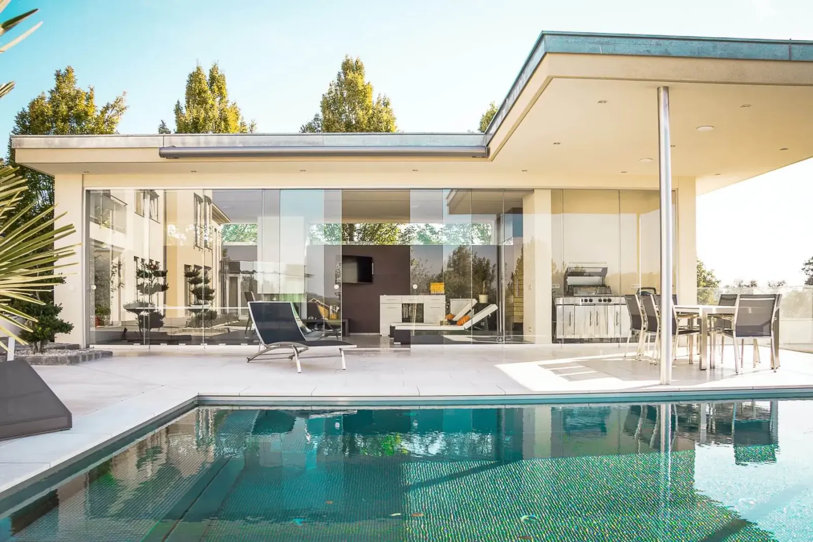 Modern luxury home with large glass walls, outdoor lounge chairs, and turquoise pool under sunny sky.
