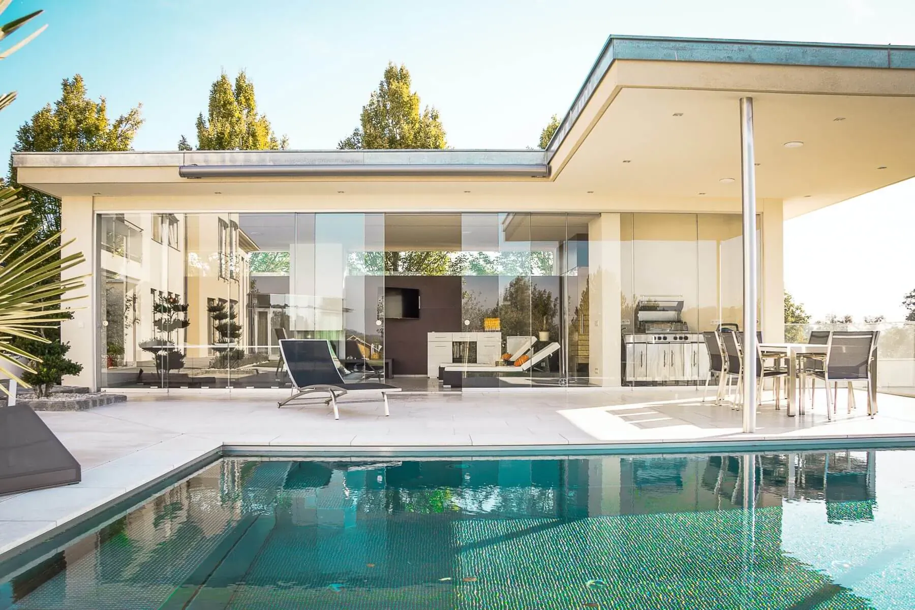 Modern luxury home with large glass walls, outdoor lounge chairs, and turquoise pool under sunny sky.