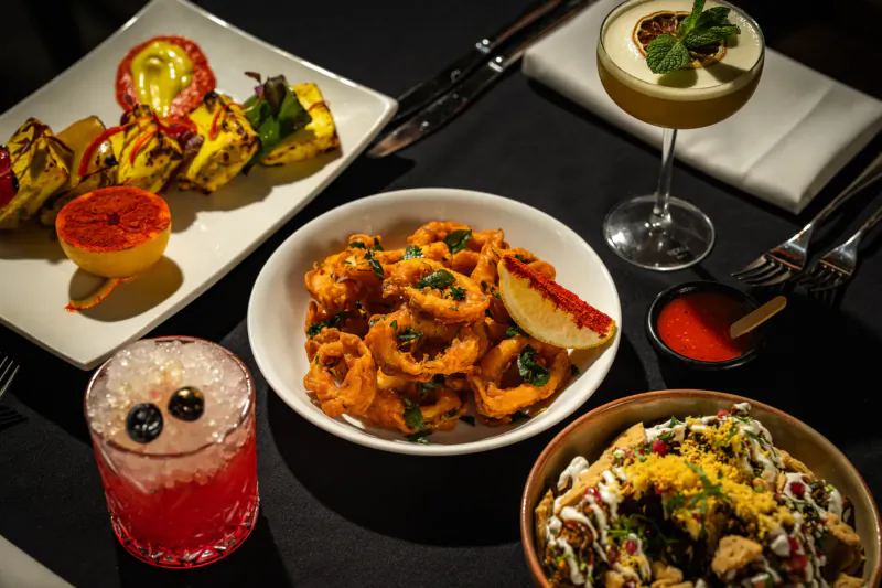 Indian dishes on black table: tandoori kebabs, calamari, chaat, with cocktails and mint garnish at Mint Leaf London.