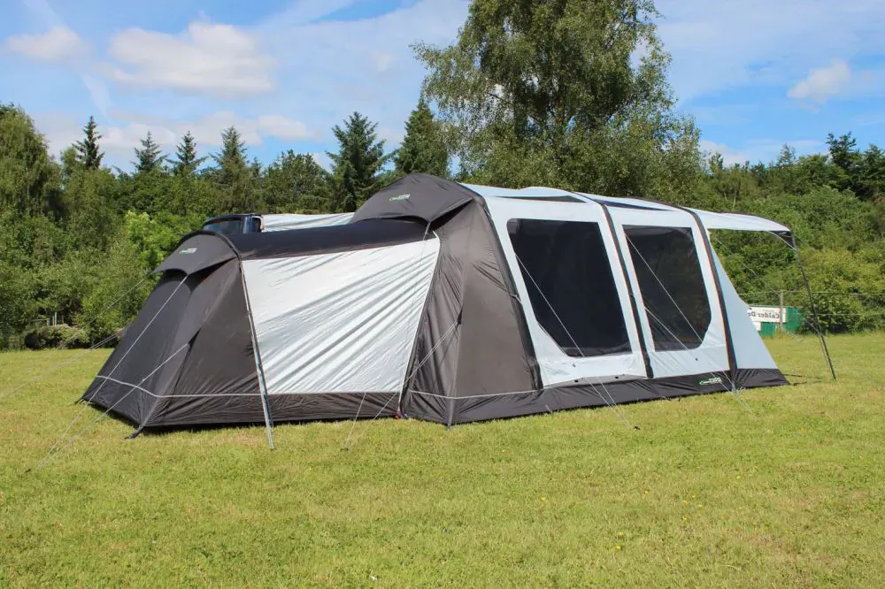 Large black and white Outdoor Revolution tent pitched on grassy field amid pine trees under blue sky.