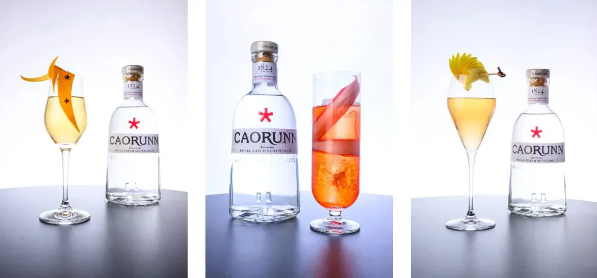Caorunn gin bottles with orange and lemon garnishes, served in glasses with fruit slices on dark surface.