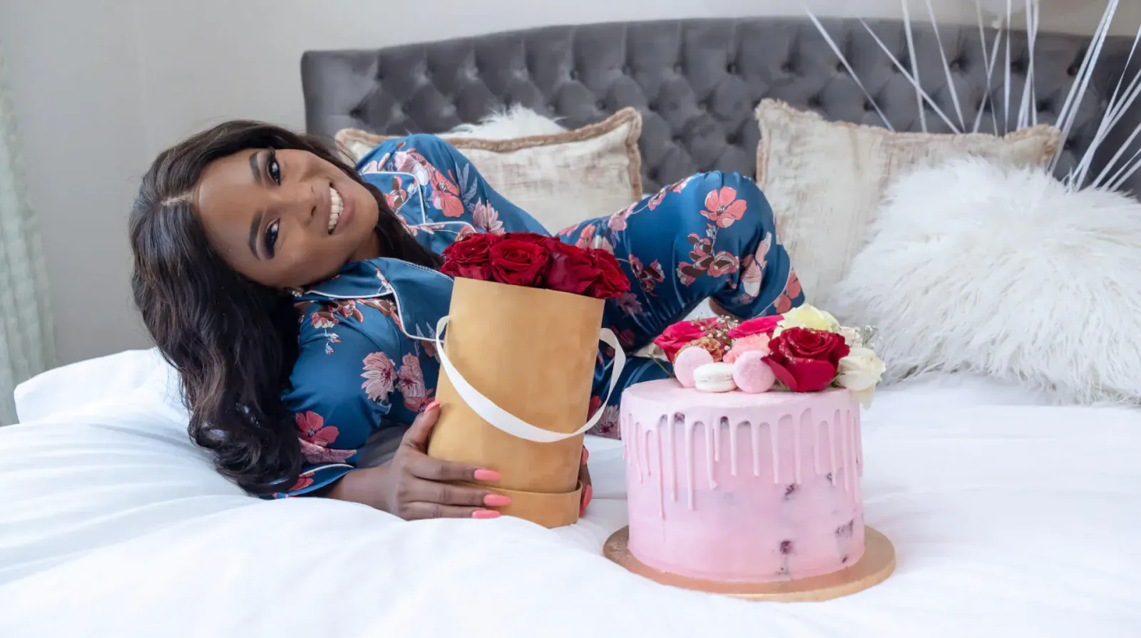 Smiling Black woman in blue pajamas lounging on bed, holding red roses box and pink drip cake.