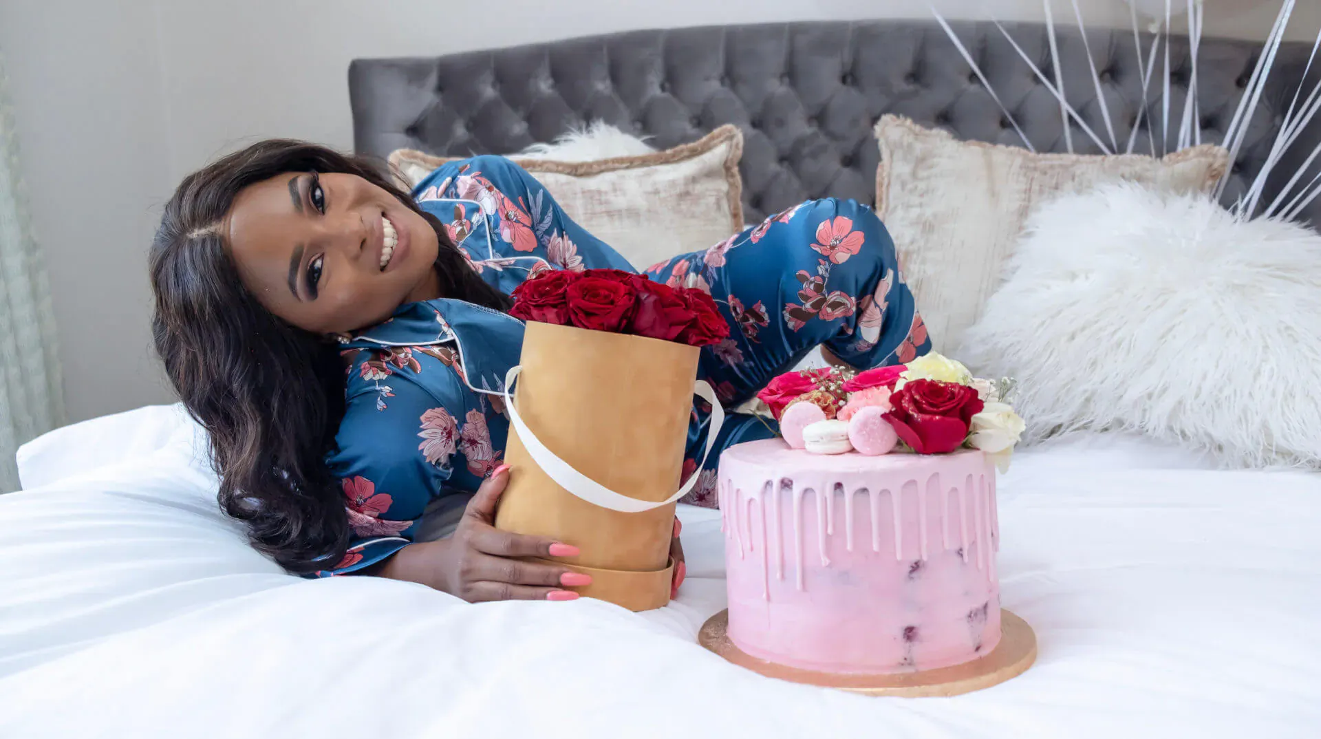 Smiling Black woman in blue pajamas lounging on bed, holding red roses box and pink drip cake.