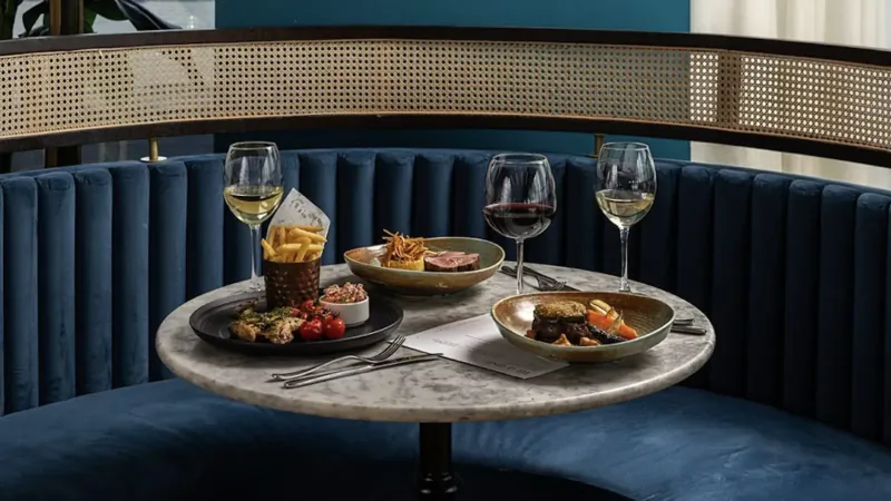 Elegant restaurant table on blue velvet booth with white wine glasses, fries in cup, veggie plates, and gold bowls.