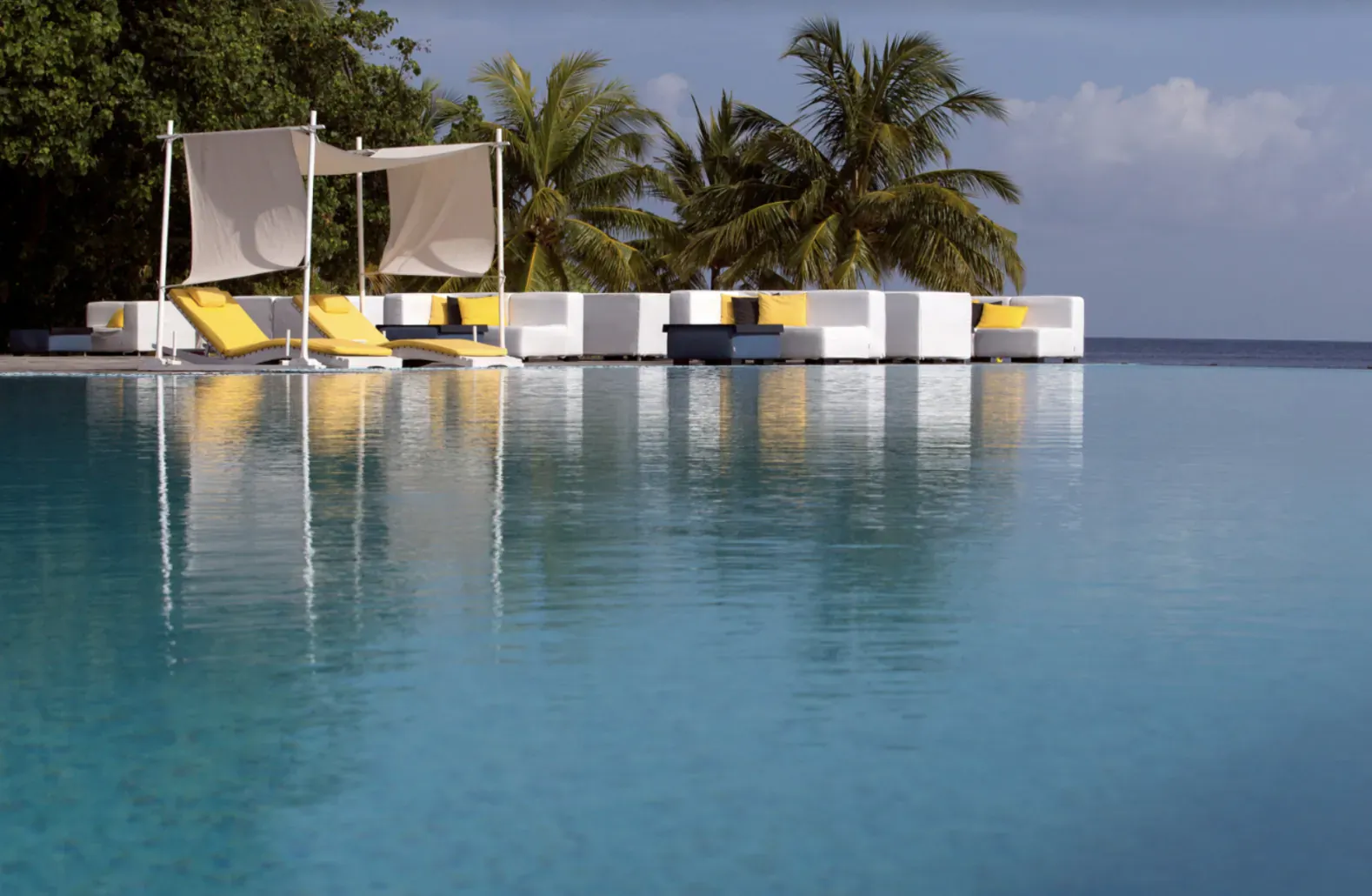 Infinity pool at Coco Bodu Hithi resort in Maldives, with white loungers under yellow umbrellas, palm trees, and ocean horizon.