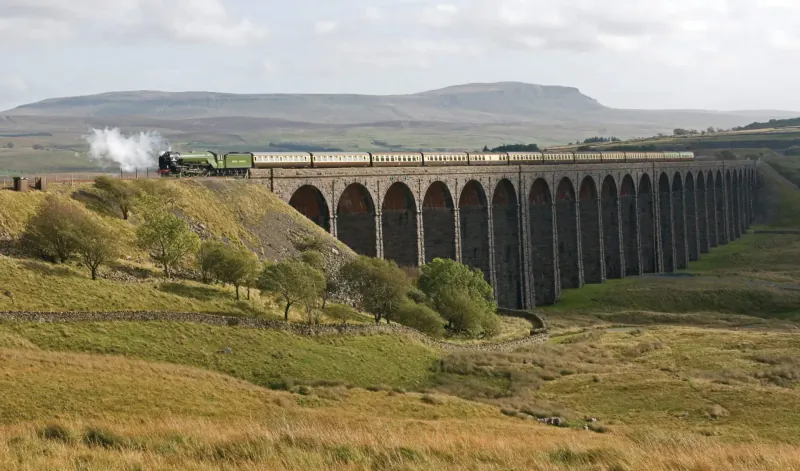 Green steam train with white steam crossing tall stone viaduct in Yorkshire moorlands under cloudy sky