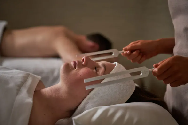 Therapist holding tuning forks to face of woman lying on spa bed with white head wrap, man relaxing nearby.
