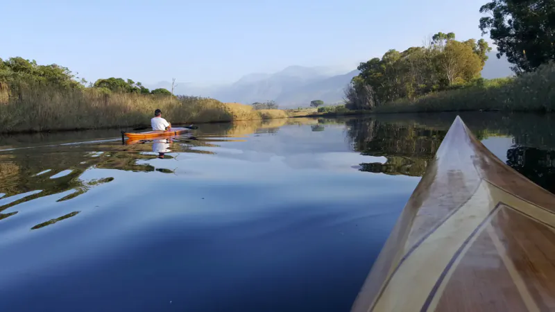 Close-up view from wooden kayak bow paddling down calm Klein River, kayaker ahead amid reeds, mountains beyond.