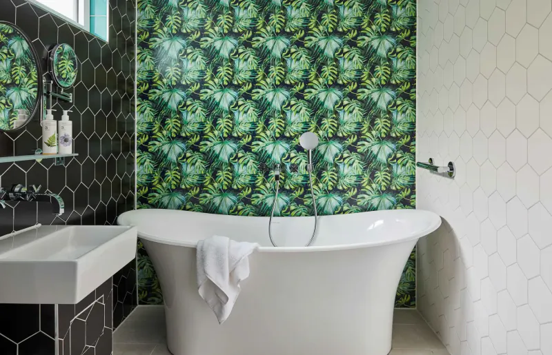 Elegant bathroom in Hotel Indigo Bath with freestanding tub draped in towel, green leaf wallpaper, black hex tiles, and rain shower.