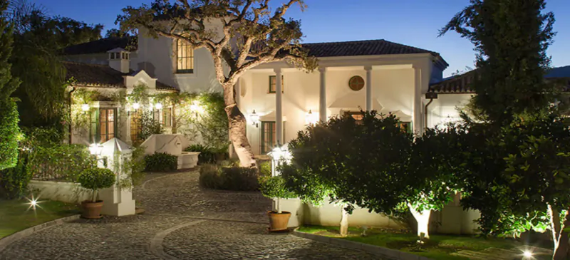 Luxury white villa at Marbella Mountain Resorts, lit at dusk with palm trees, gardens, and stone driveway.