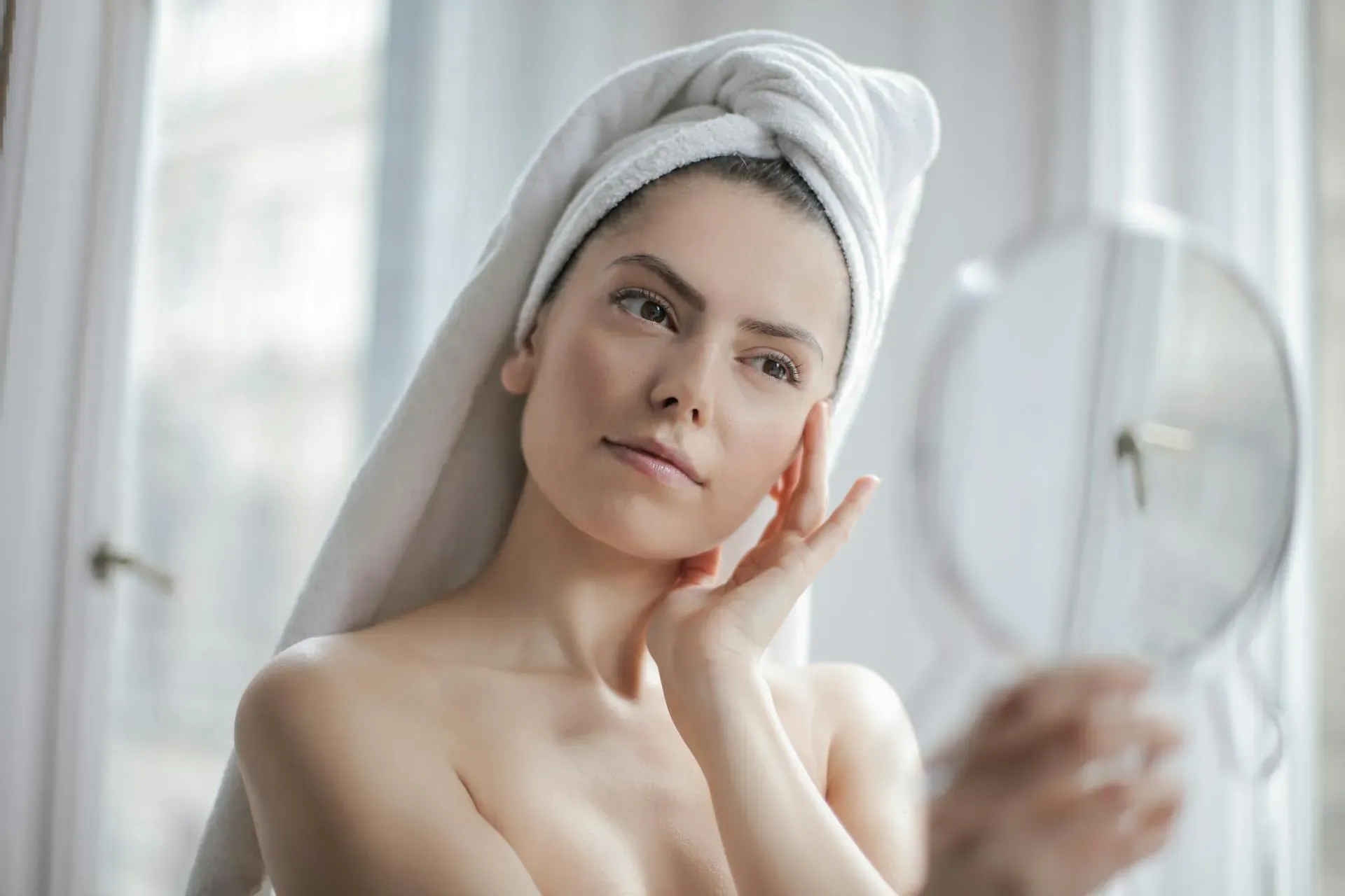 Woman with white towel on head, shirtless, touching cheek thoughtfully while holding mirror by window