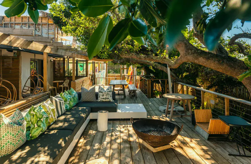 Seaside bungalow deck with lush green trees, L-shaped sofa, dining table, fire pit, and ocean view.