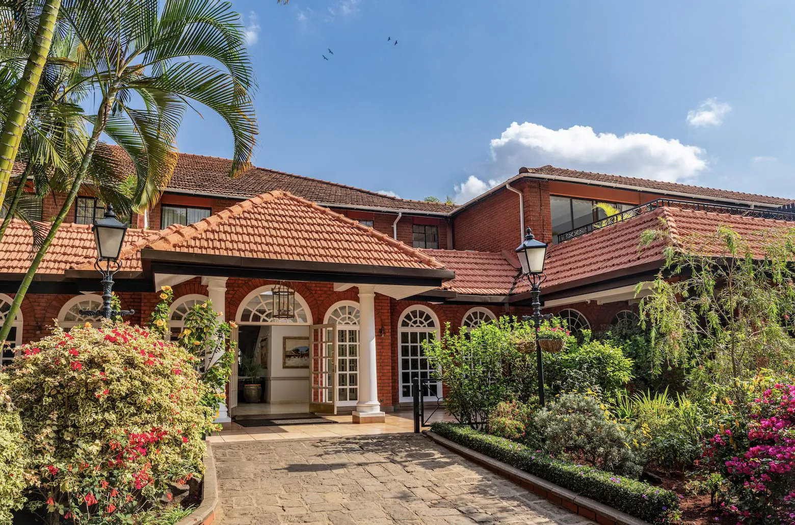 Fairmont The Norfolk hotel in Nairobi: red-tiled roof mansion with arched entrance, palm trees, bougainvillea, lanterns under blue sky.