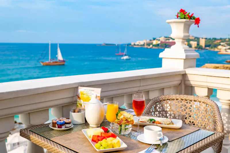 Luxury balcony breakfast table with fresh fruit, juice, croissant, overlooking blue sea, sailboats, and coastal cityscape.