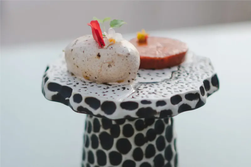 Fine dining plate on black polka-dot pedestal: white mousse topped with red leaf and microgreens, red disc beside it.