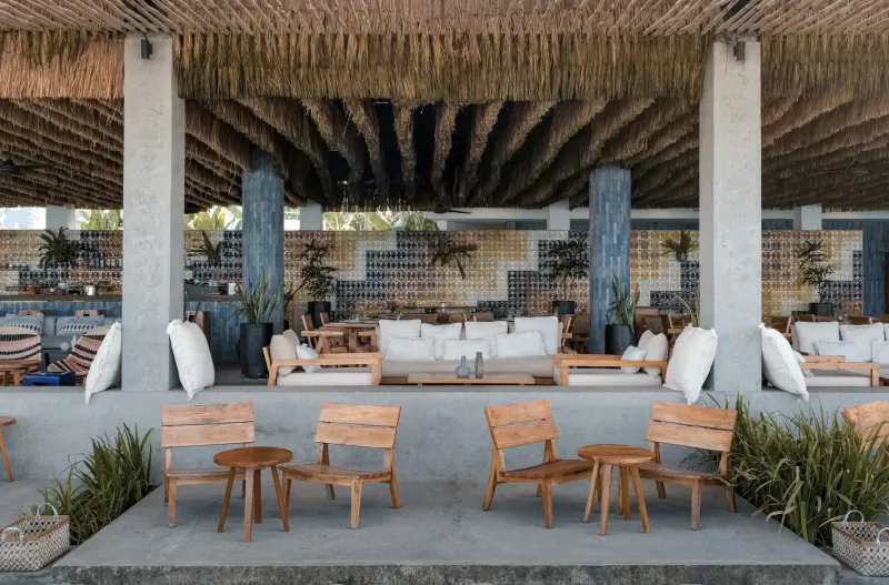 Open-air dining area at Amber Lombok with white sofas, wooden tables, plants, thatched roof, and ocean views.