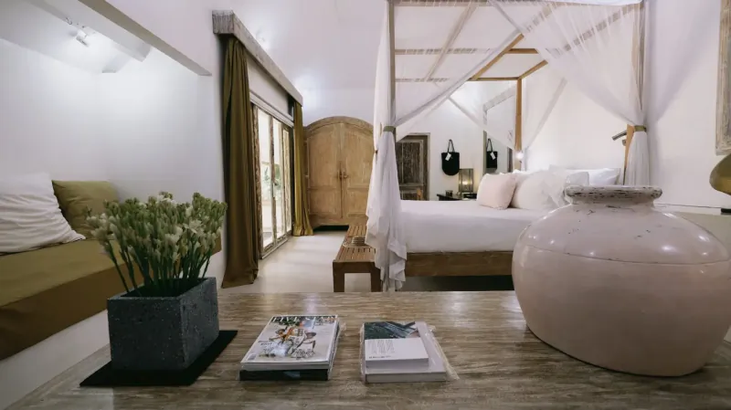 Luxurious Bali bedroom with white canopy bed, green plants, books on wooden table, and sliding doors to garden.