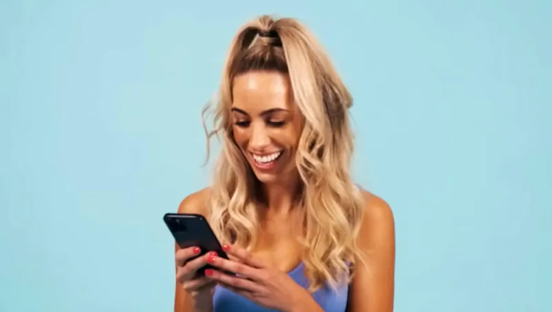 Blonde woman with ponytail in blue top smiling happily while viewing phone on blue background