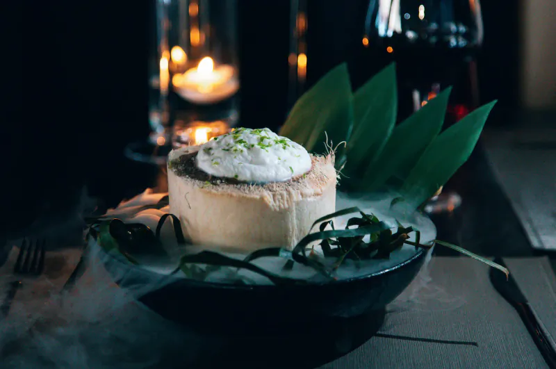 Coconut dessert topped with white cream and lime zest, garnished with green leaves in black bowl, lit by candles at Lio Ibiza.