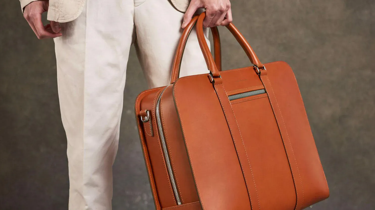 Man in white pants holding tan leather briefcase with silver accents against gray background