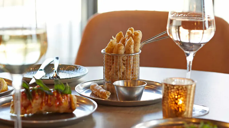 Close-up of restaurant table with fried asparagus in metal basket, spring rolls, white wine glass, and candle.