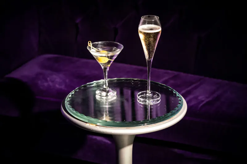 Martini with lemon twist and champagne flute on glass table against purple velvet sofa in luxury setting