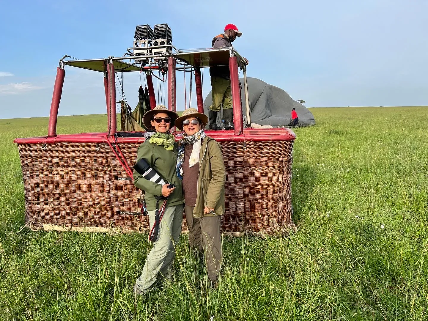 Phera and Pana in safari outfits pose smiling by red hot air balloon on grassy savanna, man in red cap nearby.