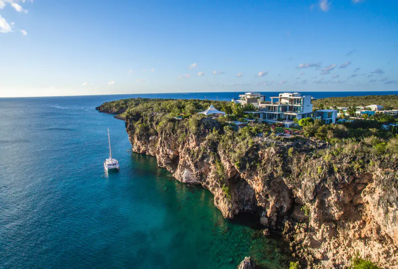 Aerial view of Àni Private Resorts: modern white villas on lush cliffs overlooking turquoise sea with catamaran below