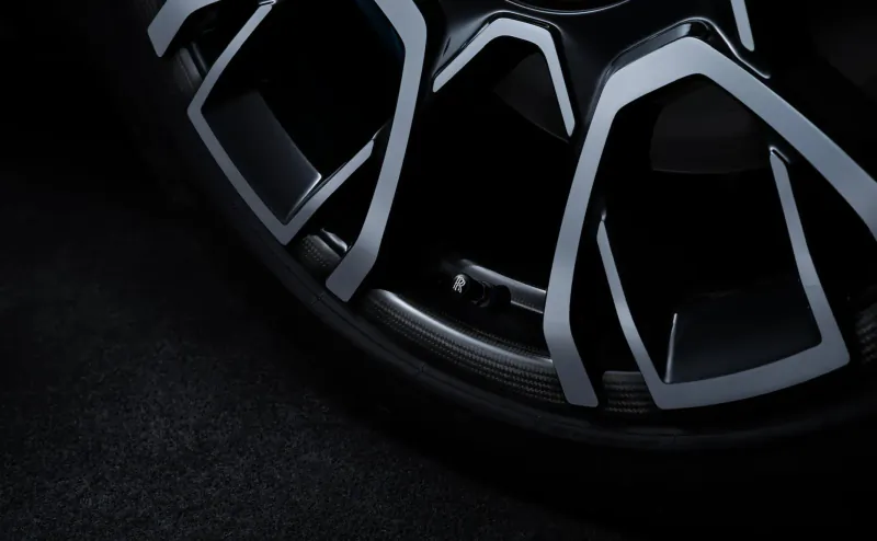Close-up of sleek black Rolls-Royce Black Badge Ghost alloy wheel with intricate angular spokes, dark studio lighting.