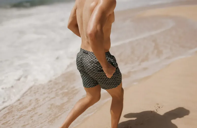 Fit shirtless man in patterned swim shorts jogging on beach by ocean waves