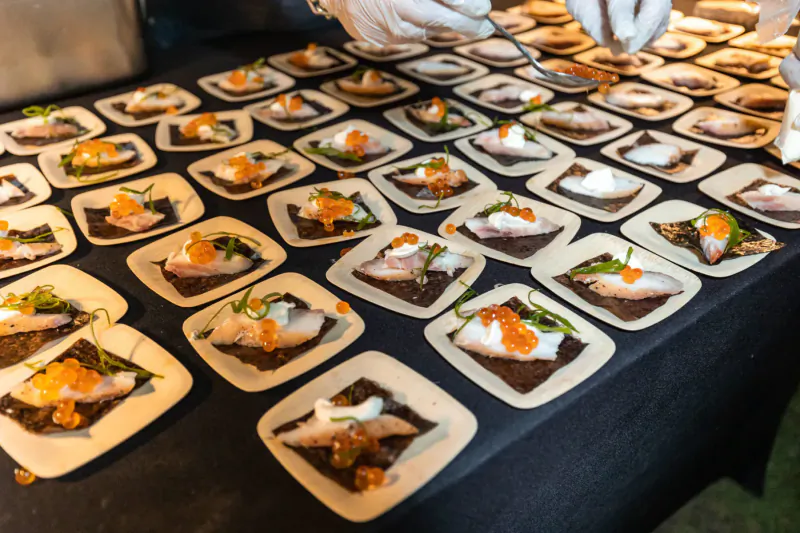 Gloved hands serving rows of plated smoked salmon with orange roe, cream, and greens at outdoor BBQ buffet.